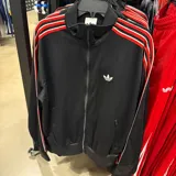 Campera deportiva Adidas Originals de color negro con cierre frontal. Presenta el logo del trébol bordado en blanco en el pecho y las clásicas tres tiras en color rojo y blanco a lo largo de las mangas. Cuenta con bolsillos laterales con cierre y puños y cintura acanalados.