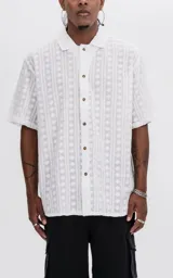 Camisa de manga corta color blanco off-white, con cuello clásico y cierre frontal de botones. Presenta un diseño con textura de rayas verticales y motivos florales bordados en toda la prenda.