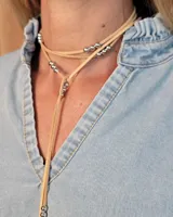 Collar tipo corbatín confeccionado en tiento de cuero, con diseño envolvente y detalles de cuentas metálicas plateadas a lo largo de la tira.