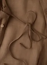 Blusa de ramio color marrón, de diseño recto, con cuello camisero, manga larga con puños abotonados, cierre frontal con botones y detalle de lazo cruzado.