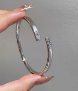 Pulsera tipo brazalete abierto de metal plateado, con un diseño ligeramente torcido en la parte inferior. Los extremos abiertos están adornados con pequeñas incrustaciones de cristales en forma de estrella, una de color rosado pálido y la otra azul claro.