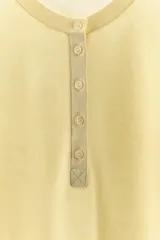 Camiseta de punto en mezcla de algodón color amarillo pastel, con cuello redondo, manga corta y cierre frontal parcial con botones.