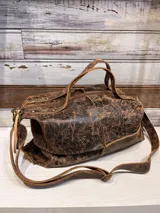 Bolso de mano tipo bowling con acabado de cuero envejecido en tonos marrones y negros. Cuenta con doble asa corta para llevar al hombro y una correa larga ajustable y extraíble para usar como bandolera. El compartimento principal tiene cierre superior y detalles de remaches metálicos.