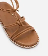 Sandalia plana color marrón con tiras finas que se atan al tobillo y suela con pespuntes en contraste.