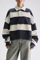 Buzo estilo rugby oversize con diseño a rayas horizontales anchas en azul marino y blanco. Presenta cuello camisero blanco con cartera de botones oculta, mangas largas y costuras expuestas con efecto deshilachado.