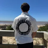 Camisa de lino color blanco con corte boxy, manga corta y cuello clásico. Presenta un estampado gráfico circular en color negro en la espalda y detalle de etiqueta pequeña en la parte superior.