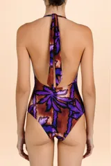 Traje de baño enterizo con diseño halter, escote profundo y estampado botánico en tonos púrpura y marrón cobrizo. Presenta una faja ancha fruncida en la cintura con un lazo decorativo central que cae hacia la parte inferior.