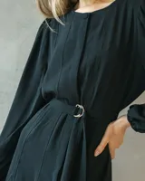 Vestido midi negro de silueta fluida, con mangas largas abullonadas y cintura marcada mediante un cinturón incorporado con hebilla metálica. Presenta un diseño de escote cerrado y caída liviana.