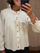 Camisa de algodón color blanco con cuello redondo y detalle de fruncido tipo nido de abeja en el canesú. Presenta cierre frontal con botones decorativos, mangas largas abullonadas con puños elásticos fruncidos y volados en el cuello.