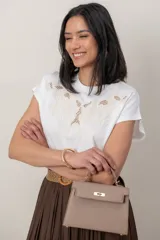Cartera de mano tipo satchel de eco cuero color beige topo, con herrajes metálicos dorados, solapa frontal con cierre de broche giratorio y asa corta superior. Incluye correa para colgar.