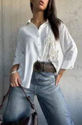 Camisa blanca de corte holgado con cuello clásico, cierre frontal de botones y detalle de bordado en relieve en el frente. Posee mangas largas con puños doblados.