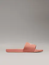 Ojotas tipo slide de color rosado, con base de EVA y correa ancha de lona de algodón reciclado. La correa presenta el logo de Calvin Klein Jeans bordado en blanco.