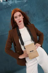 Bandolera pequeña tipo porta celular, color beige, confeccionada en cuero sintético. Cuenta con una solapa frontal con cierre magnético y detalle de etiqueta con logo de la marca. Incluye una correa de hombro ancha, ajustable y extraíble, con diseño de letras grabadas.
