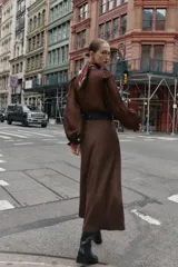 Vestido midi de color marrón con diseño combinado, escote en pico con volantes, mangas largas con puños elásticos y falda evasé. Incluye un cinturón negro con hebilla dorada en la cintura y cierre lateral con cremallera oculta.