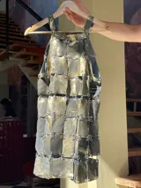 Vestido corto sin mangas, con tirantes finos, confeccionado artesanalmente con piezas rectangulares de cuero genuino unidas por herrajes plateados. El cuero tiene un acabado pintado a mano en tonos gris oscuro y dorado brillante.