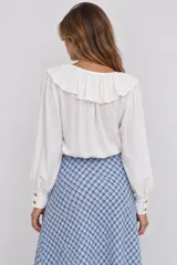 Blusa blanca de manga larga con escote en V y cuello con volados. Cuenta con lazos para atar en el pecho y puños anchos con botones dorados.