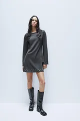 Vestido corto de punto color gris oscuro, con cuello barco y mangas largas acampanadas. Presenta detalles de tachas metálicas aplicadas en el ruedo y en los puños, con un diseño de corte evasé.