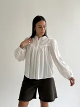 Camisa blanca de algodón con cuello mao, mangas largas con puños elásticos y cierre frontal de botones. Presenta un diseño con pliegues en la parte delantera y detalles bordados en el pecho.