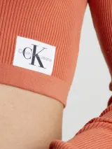 Sweater de punto acanalado color terracota, de manga larga y corte cropped ajustado. Presenta un cuello de chomba con escote en pico profundo y una pequeña etiqueta con el monograma de Calvin Klein en el bajo.