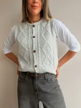 Chaleco tejido de punto en color gris claro, con diseño de trama de rombos y trenzas en el frente. Presenta cuello redondo, cierre frontal con botones oscuros y terminaciones en punto elástico.