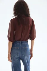 Blusa de tejido liviano color marrón oscuro con efecto arrugado. Presenta un corte holgado, cuello elevado, botones forrados en el frente y canesú fruncido de doble capa. Las mangas son globo a tres cuartos con puños ribeteados con cinta fina.