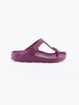 Sandalias tipo ojota de goma, con diseño de dedo y tira ajustable con hebilla lateral. Presentan una plantilla anatómica y un pequeño aplique circular decorativo en la parte frontal.