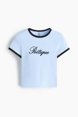 Remera corta celeste entallada de algodón, con cuello redondo y ribetes en contraste negros en el cuello y las mangas. Cuenta con un estampado frontal con la palabra "Poétique" en cursiva.