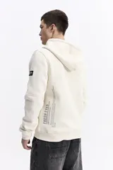 Campera con capucha y cierre frontal, color crema, con bolsillo canguro. Presenta un estampado de texto en el pecho y detalles en el puño.