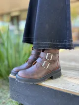 Botas cortas de cuero color marrón con suela track negra. Presentan dos correas ajustables con hebillas metálicas plateadas en el empeine y cierre lateral interno.