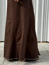 Pollera larga de tiro alto confeccionada en denim color marrón, con diseño ajustado en la parte superior y corte acampanado hacia el ruedo. Presenta costuras verticales decorativas y un acabado desflecado en el borde inferior.