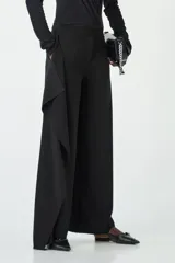 Pantalón negro de tiro bajo, corte ancho y recto, con un detalle de tela superpuesta en forma de bolado o drapeado asimétrico en la parte frontal de una de las piernas.