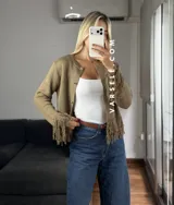 Cárdigan de tejido de punto color crema, con cuello redondo, mangas largas y cierre frontal con botones metálicos. Presenta detalles de flecos en los puños y en el ruedo.