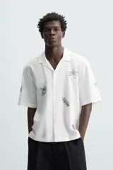 Camisa de manga corta confeccionada en mezcla de algodón y lino, color blanco. Presenta un cuello tipo bowling, calce regular y cierre frontal con botones. Destaca por sus bordados lineales en contraste de color negro con motivos de frutas y hojas distribuidos por toda la prenda.