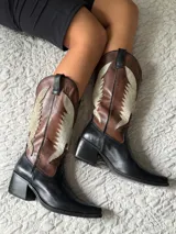 Botas texanas de caña media confeccionadas en cuero, con diseño bicolor que combina negro en la base y puntera, y marrón con detalles en cuero texturizado brillante en la caña. Presentan un taco cuadrado de 5 cm y diseño sin cierre.