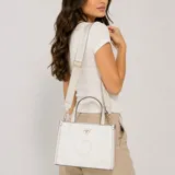 Cartera tipo tote Guess modelo Jackie, de color blanco crema con estampa de monograma y textura granulada. Cuenta con doble asa de mano, correa ajustable de tela, herrajes dorados y el logo triangular característico de la marca en el frente.