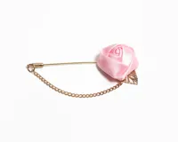 Pin para el ojal con diseño de rosa de satén rosado, acompañado de una hoja metálica dorada y una cadena de eslabones fina que conecta ambos extremos del alfiler.