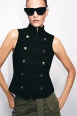 Chaleco negro de cuello subido y manga sisa, con cierre frontal oculto y detalle de botones decorativos metálicos en estilo militar.