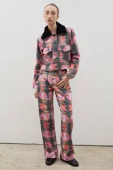 Pantalón de tiro alto confeccionado en denim de corte recto, con estampado artístico abstracto en tonos rosados, rojos y negros. Presenta cierre frontal con botones visibles.