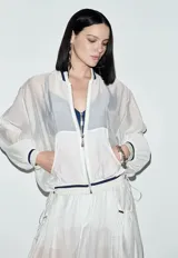 Chaqueta bomber blanca transparente con cierre frontal de cremallera, cuello, puños y cintura elásticos a rayas azul marino, bolsillos laterales y mangas abullonadas.