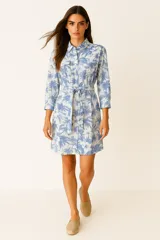 Vestido camisero corto de algodón con estampado botánico en tonos azules y blancos. Presenta cuello camisero, cierre frontal con botones, mangas tres cuartos y bolsillos frontales con solapa. Incluye un lazo ajustable en la cintura para entallar la silueta.
