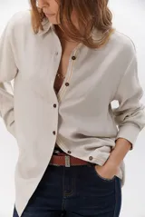 Camisa beige de manga larga con botones marrones y cuello clásico. Presenta un detalle de costura dorada a lo largo de la botonera.