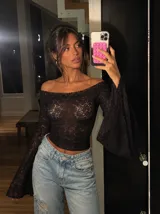 Blusa de encaje negro con diseño de hombros descubiertos y mangas largas acampanadas. Presenta un ajuste al cuerpo y tejido transparente.