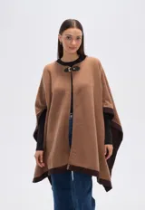 Poncho de tejido grueso color camel, con bordes y mangas en color marrón oscuro. Cierre frontal con broche rectangular negro.