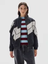 Campera de denim estilo bomber en color negro lavado, con paneles en contraste color crema formando un diseño en zigzag. Presenta cierre frontal, cuello alto y puños elásticos.