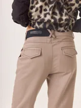 Pantalón color beige, corte tipo cigarette con pierna angosta. Presenta costuras contrastantes en color negro y bolsillos traseros con tapa. La cintura es alta y tiene un detalle de cruce en la espalda con un parche de cuero negro con logo grabado.