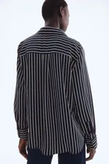 Camisa de manga larga con cuello clásico y cierre frontal de botones. Presenta un estampado de rayas verticales finas blancas sobre fondo azul marino. Los puños son anchos y también tienen rayas horizontales.