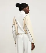 Campera deportiva de cuello alto con cierre frontal completo. Presenta un diseño bicolor en blanco y beige con líneas curvas en negro que definen la silueta. Confeccionada en una mezcla de poliéster, modal y elastano.