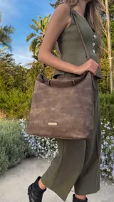 Cartera tipo bandolera de gran tamaño, confeccionada en cuero sintético con acabado desgastado. Cuenta con una asa corta fija, una correa larga regulable y extraíble, y una placa metálica con la marca en la parte frontal.