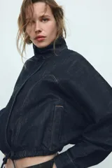 Cazadora de denim color azul oscuro, con cuello solapa, mangas largas tipo murciélago y bolsillos de vivo en el delantero. Presenta un bajo elástico ajustable con cordones y cierre frontal con cremallera y botones a presión ocultos.