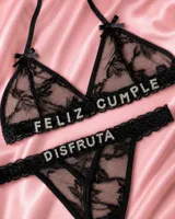 Conjunto de lencería de dos piezas confeccionado en encaje negro, compuesto por un soutiene triangular con tirantes halter y una bombacha tipo colaless. Presenta aplicaciones de letras brillantes con pedrería que forman la frase "FELIZ CUMPLE DISFRUTA".